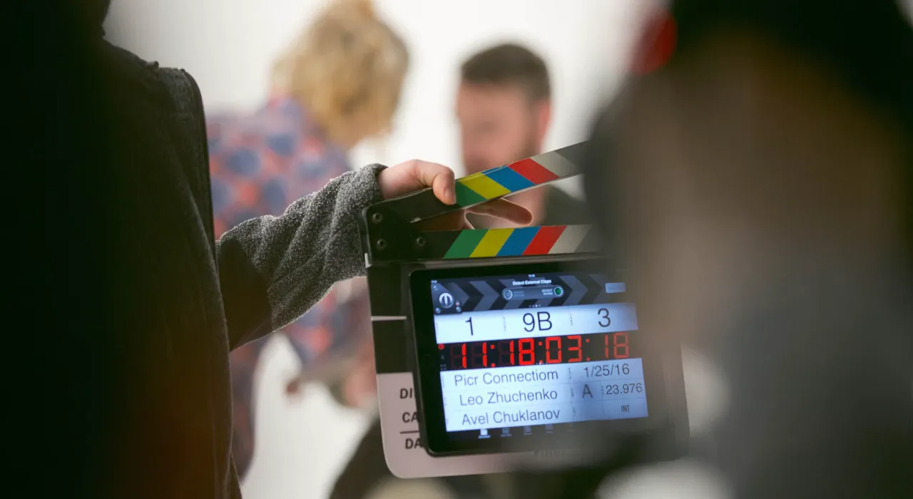 A crew member holds a digital clapperboard showing scene 9B, take 3, in front of two blurred actors in a white studio.