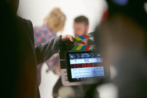 A crew member holds a digital clapperboard showing scene 9B, take 3, in front of two blurred actors in a white studio.