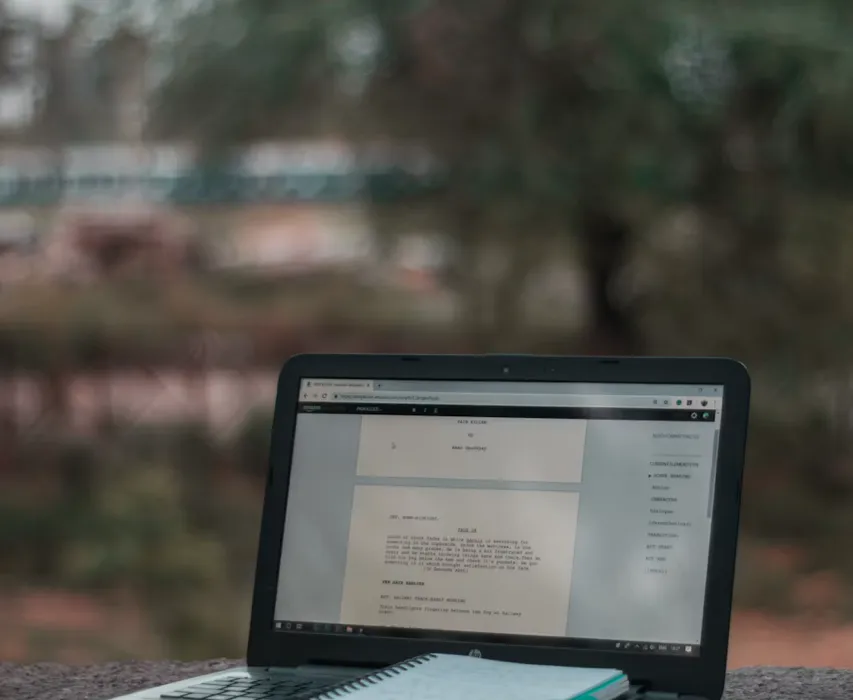 An open laptop showing screenwriting software, with a spiral notebook on the keyboard, on a stone ledge outdoors.